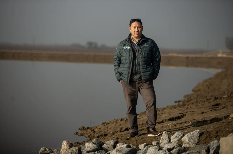 An Asian man stands near a pond wearing a green vest and dark pants.