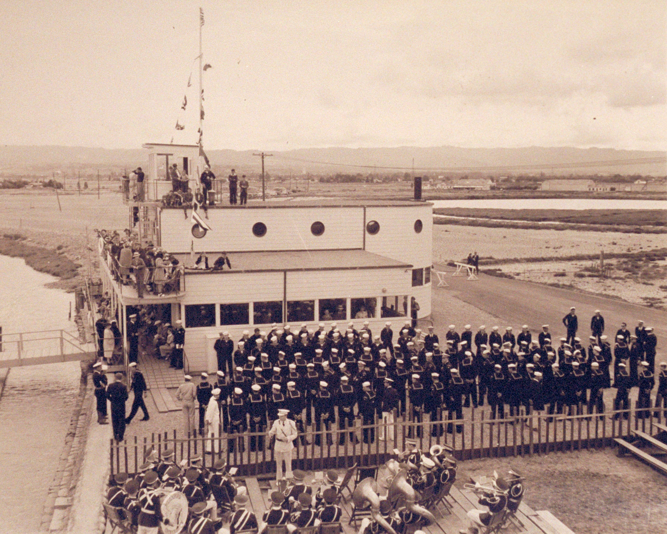 A sepia toned photograph shows rows of young men in maritime uniforms in front of an Art Deco building that looks like a boat. What looks like a military band plays in foreground.