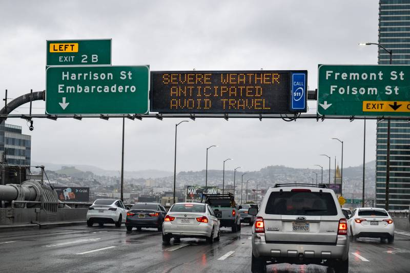 Cars head west, at the end of the Bay Bridge, toward San Francisco on a rainy day. A highway digital sign says 'Severe weather anticipated - avoid travel.'