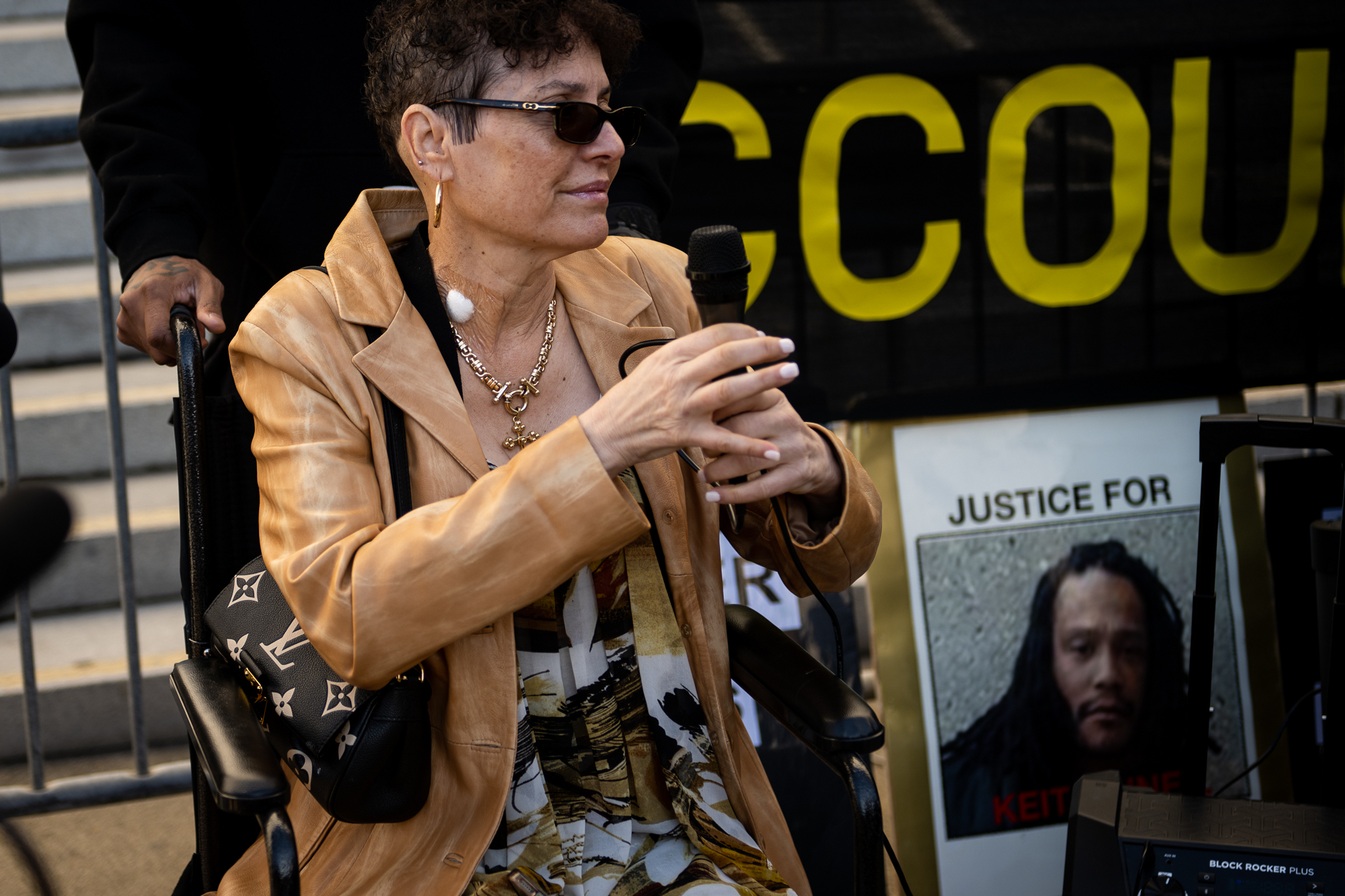 A middle-aged woman with light, freckled skin and short dark hair is seated in a wheelchair holding a microphone, next to a photo of a Black man with long black locs. The woman wears a beige leather jacket and a patterned matching blouse, with gold earrings and a necklace.