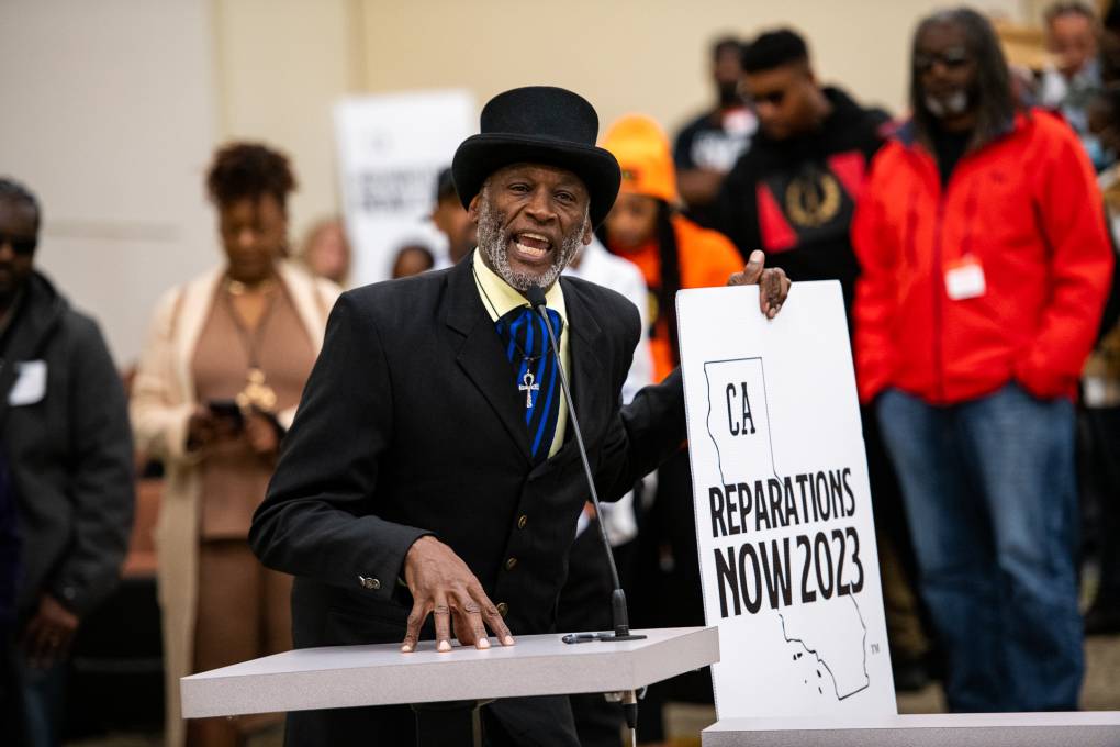 A older African American man in a black suit with a top hat holds a sign in a conference hall surrounded by people. The sign reads "CA Reparations Now 2023."