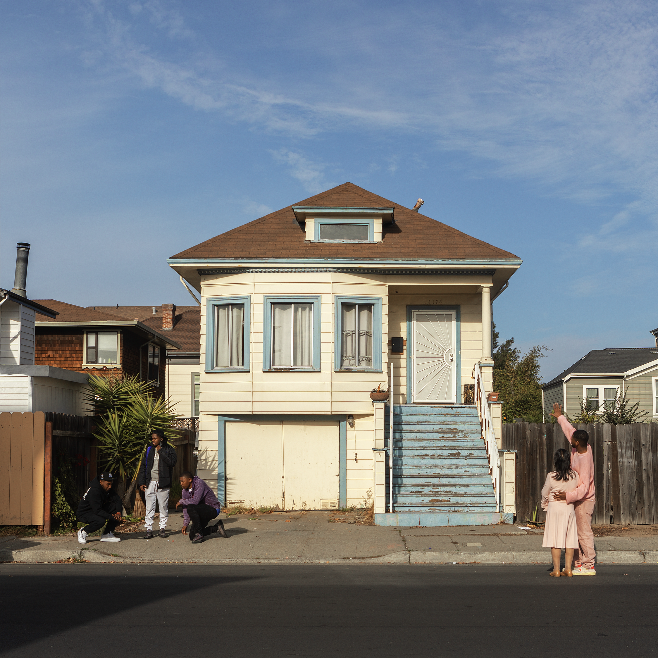 A weathered white and light blue house sits in the middle as a young man in a light pink track suit points toward it with one hand, while holding his grandmother with the other. To the left, three teenagers are gathered as two kneel on the sidewalk playing dice.
