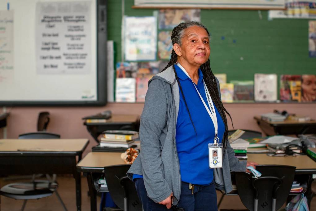 A woman with long, brown and gray braids stands tall in a classroom. She's wearing a gray, zip-up sweatshirt and a blue T-shirt. A lanyard with her name and photo lies around her neck. Rows of desks and chairs are in the background as well as chalkboards and school posters.