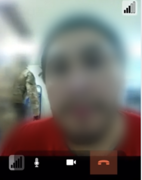 A screenshot of a video call, showing a blurred faced of a detained man, with a glimpse of a guard in military gear in the background.