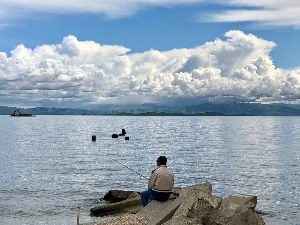 A man sits on some rocks fishing in the center of the photo. He is facing away from the camera. Beyond, a body of water stretches in front of him and a small range of hills in the far distance.