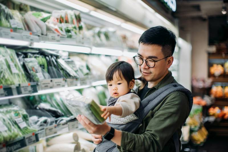 A young Asian man with short dark hair and round glasses carries a 1-year-old girl, with tiny black pigtails, in a harness on his chest, with the girl facing out. They stand in the light of a vegetable display in a supermarket. The man holds a plastic container full of green vegetables, maybe cucumbers, smiling as his daughter reaches out to touch the box.