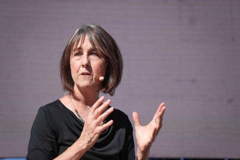 A middle aged white woman with straight grayish brown neck-length hair and a black top photographed while speaking and with hands gesturing.