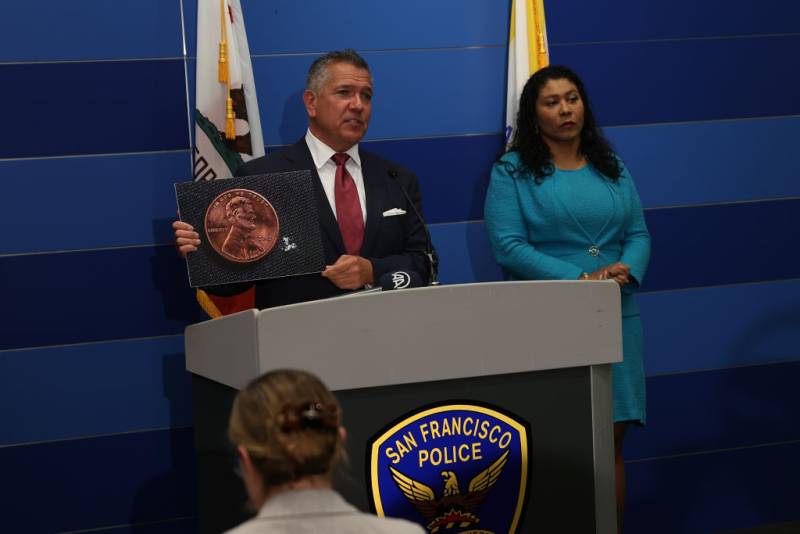 A white man in a blue suit with a red tie speaking behind a dais with a microphone and a San Francisco Police emblem with an African American woman with shoulder length hair and a blue dress listening on.