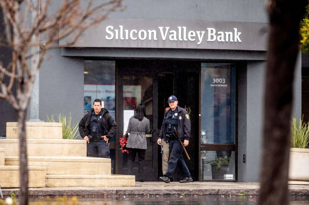 Two armed police officers, one with a baton in left hand, stand outside Silicon Valley Bank headquarters as another person behind them reads a notice taped to the closed front entrance explaining the bank's closure.