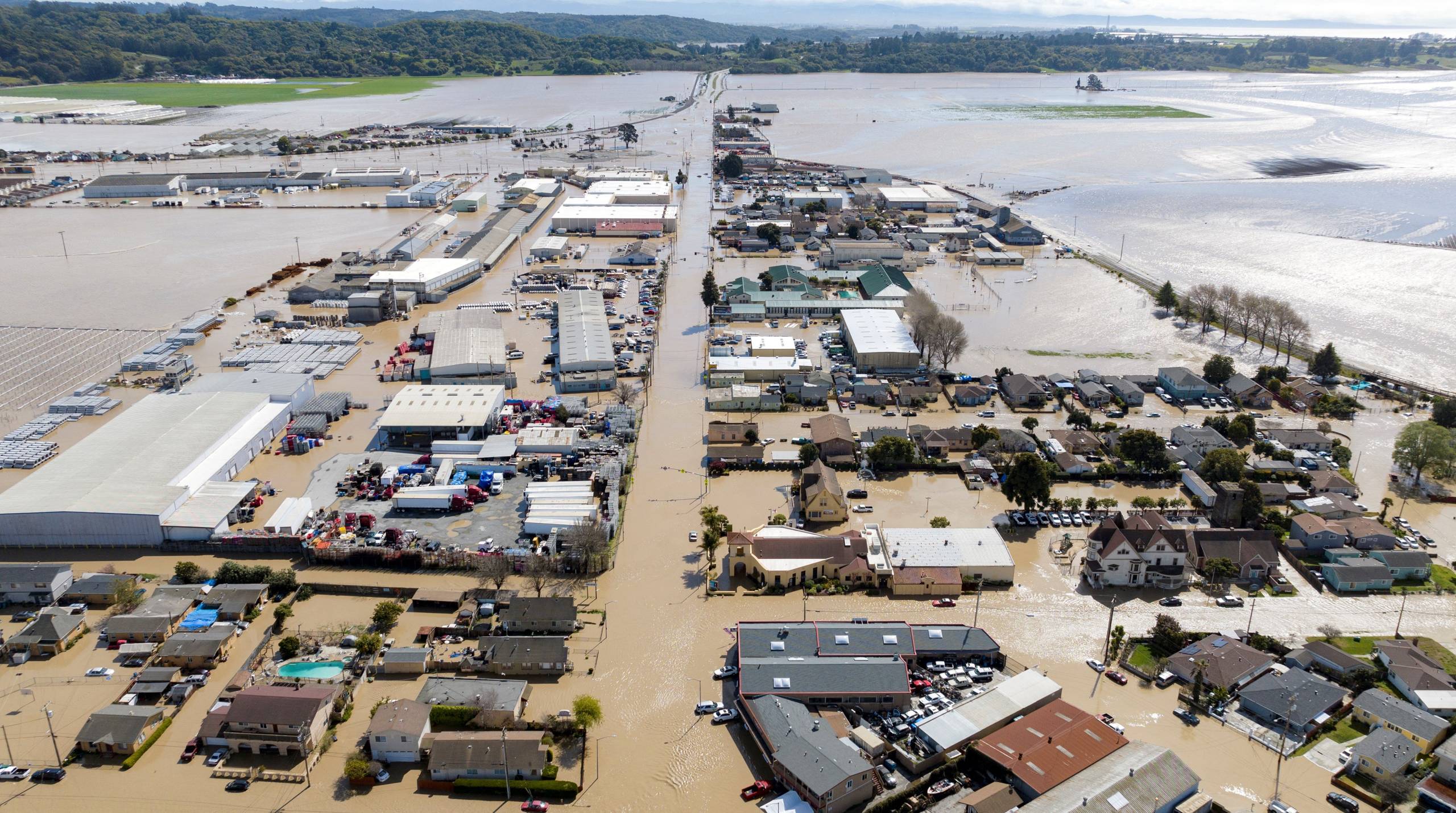 An aerial view shows many buildings, homes, streets and cars flooded with brown water.