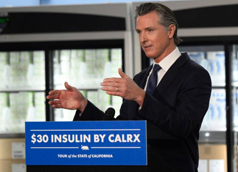 A white middle-aged man with a dark suit and tie gesticulates while speaking behind a dais on which is written "$30 insulin by CalRx" with freezers full of medicine behind him.