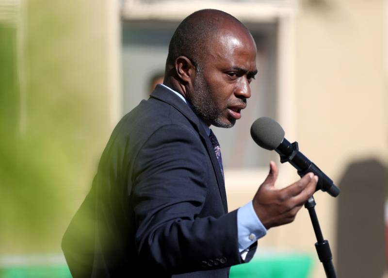 An African American man in a suit speaks into a microphone as he gestures with his hand.