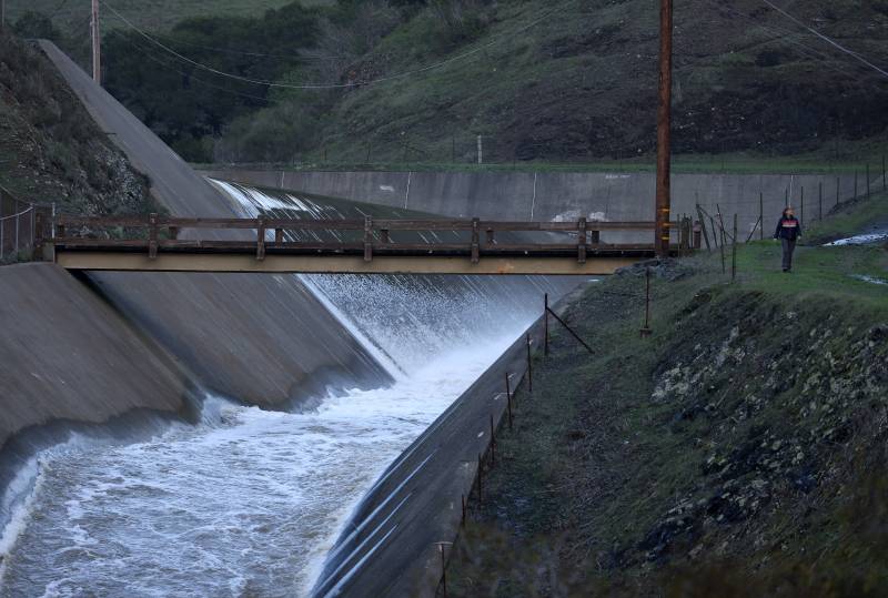 Rushing white waters rapidly flow down the spillway at the Nicasio Reservoir in California as a person in a black hoodie and jeans walks along the levee to the right.