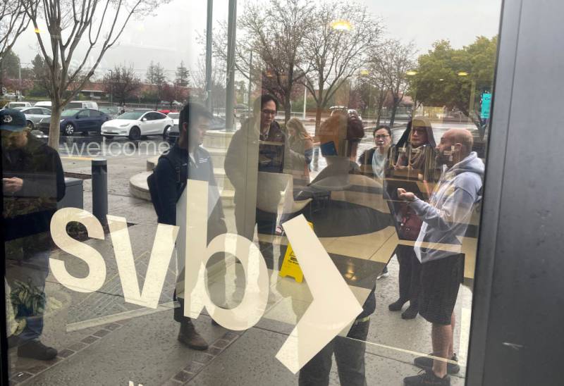 A small crowd of people is seen reflected from the window of SVBN headquarters, with the word "SVB" on the window in the foreground. Behind the crowd is a carpark on a grey and cloudy day.