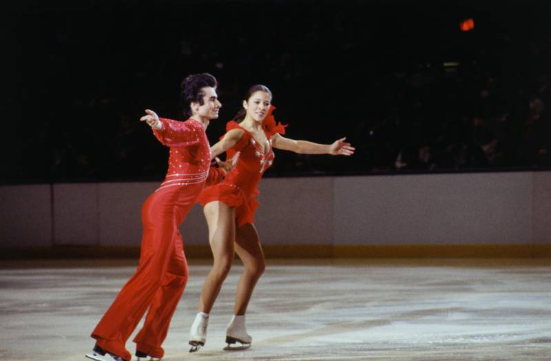 A male and female figure skater wearing matching red, sparkly outfits glide across the ice.