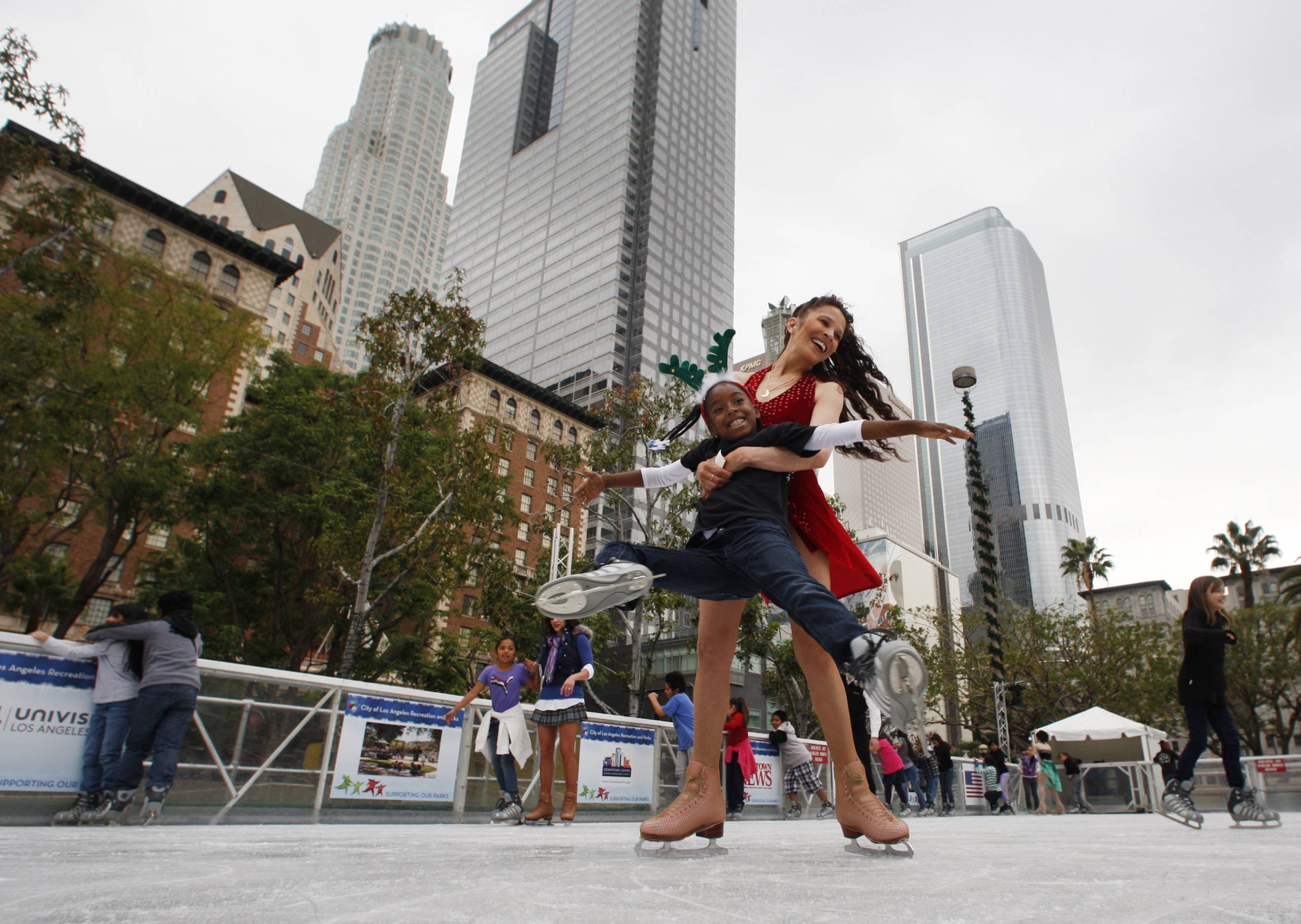A female figure skater wearing a sparkly, red outfit spins around a child wearing a navy blue school uniform as they smile together on an ice rink outdoors with buildings and skyscrapers in the background.