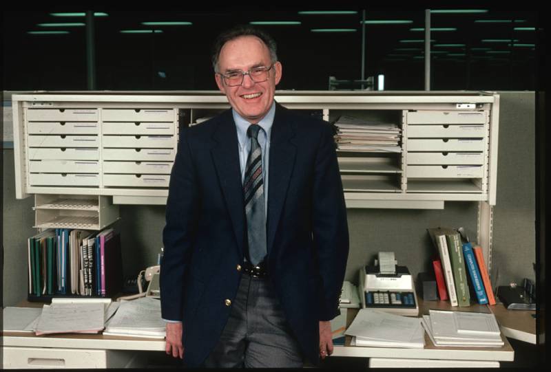 A middle-aged white man with glasses and wearing suit and tie smiles at the camera in his office.