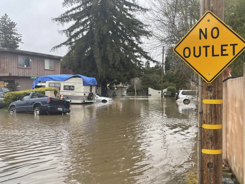 A flooded street with cars and houses and brown water, a sign that says "No Outlet"