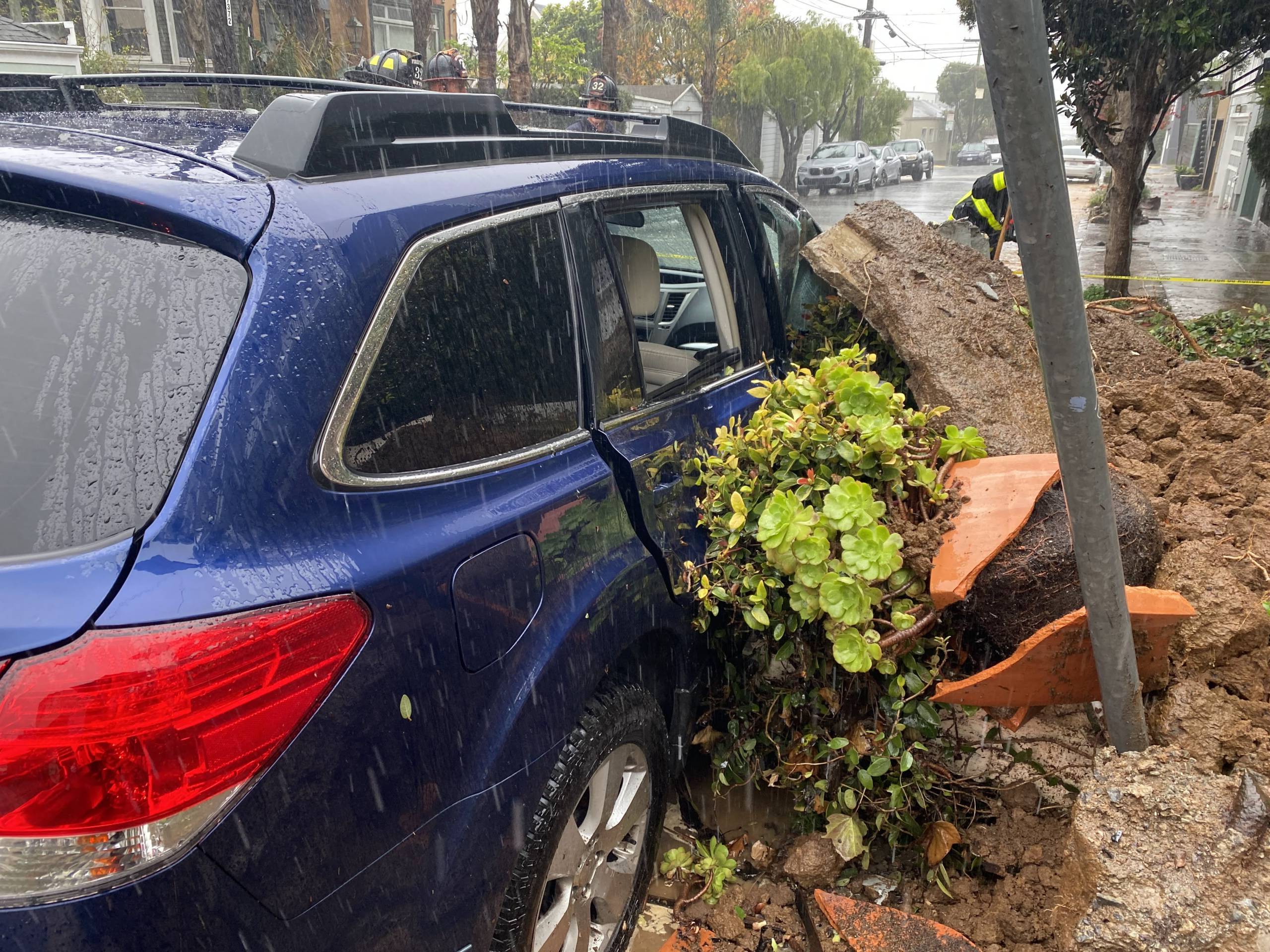 Rain pours down on a navy blue Subaru Outback that is surrounded by rubble and debris from a landslide that totaled the vehicle.