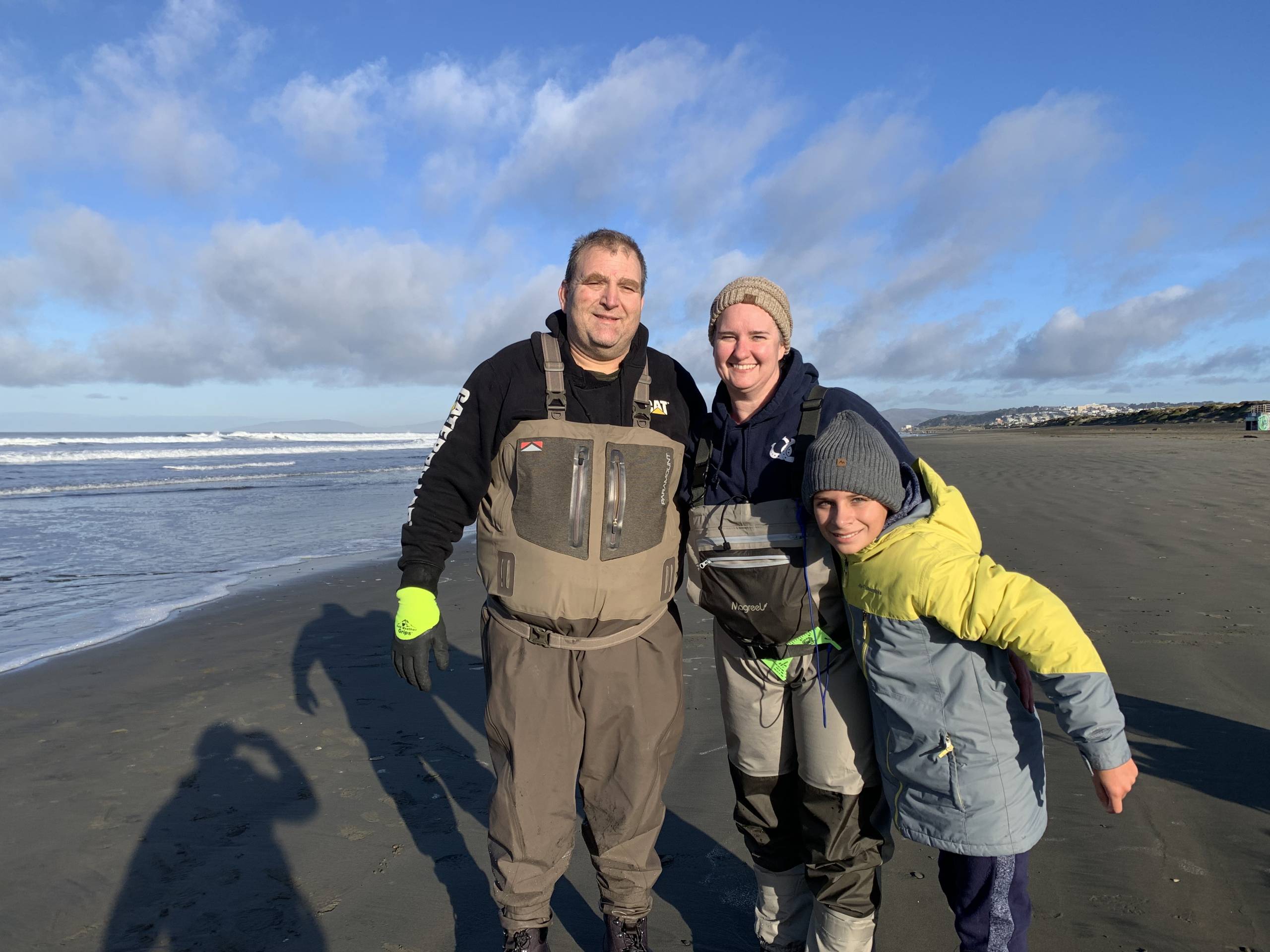 A man, woman and child smiling for the camera on a beach. They are wearing waders for fishing.