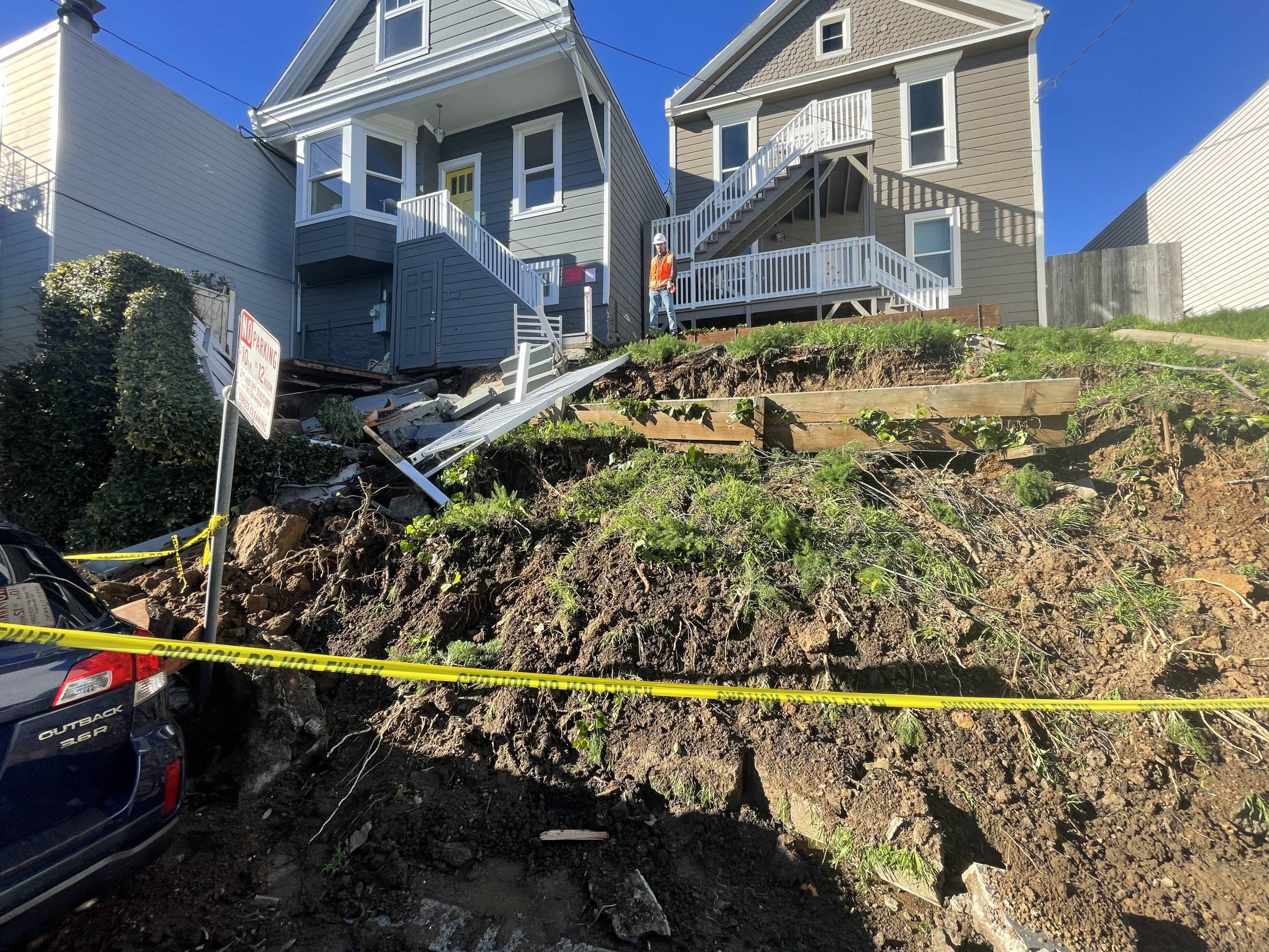 A Subaru Outback is buried beneath rubble and dirt from a landslide. One worker stands at the top of a hill with two houses behind him. Yellow caution tape blocks off the perimeter.