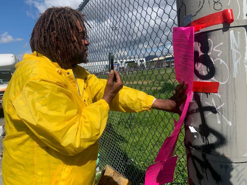 An African American man with a yellow rain jacket and dreadlocks records an eviction notice with his phone.