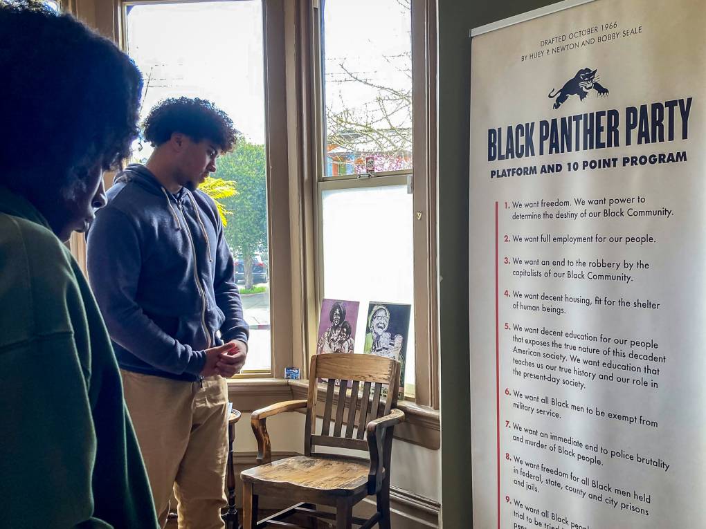 Two high school students stand next to each other inside the West Oakland Mural Project, while reading text on a wall that outlines the Black Panther Party's platform and 10-point program.