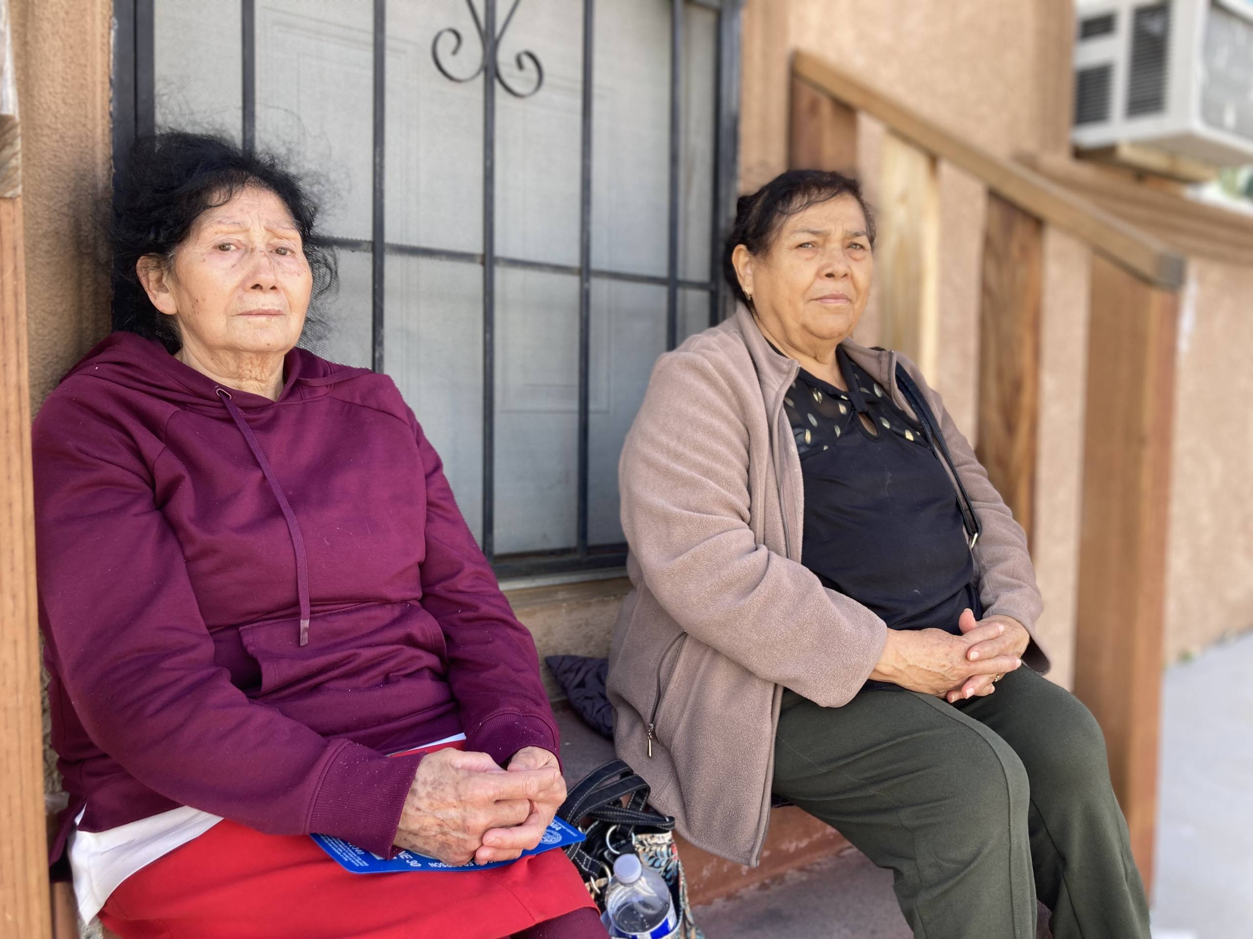 Two older women sit side by side on the steps to an apartment's front door. Each woman has her hands folded neatly on their laps. They look serious, tired, concerned.
