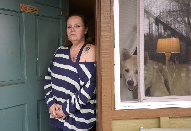 A middle-aged white woman with a long-sleeved shirt with horizontal blue-and-white stripes, with one shoulder pulled down to reveal a tattoo of an eye, looks at the camera as she leans against a door jamb. To her left, a white husky dog looks out from a window.