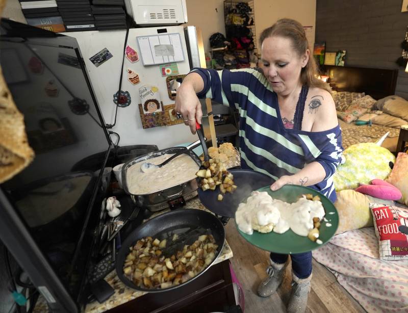 A white woman, standing in a crowded room of unmade beds, dishes potatoes onto a plate of biscuits and gravy from a hot plate on a cabinet next to a fridge. 