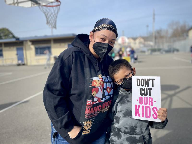 A smiling woman and child stand next to each other on a playground with their arms around each other, smiling and wearing masks. The child is holding a sign reading 'don't cut our kids'