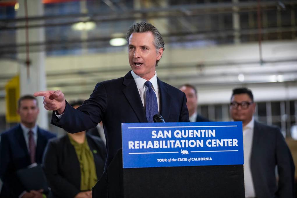 Gov. Gavin Newsom, wearing a navy suit, white-collared shirt, and blue necktie, points to his right as he speaks from a podium with a sign that reads, "San Quentin Rehabilitation Center" during his tour of the state of California.