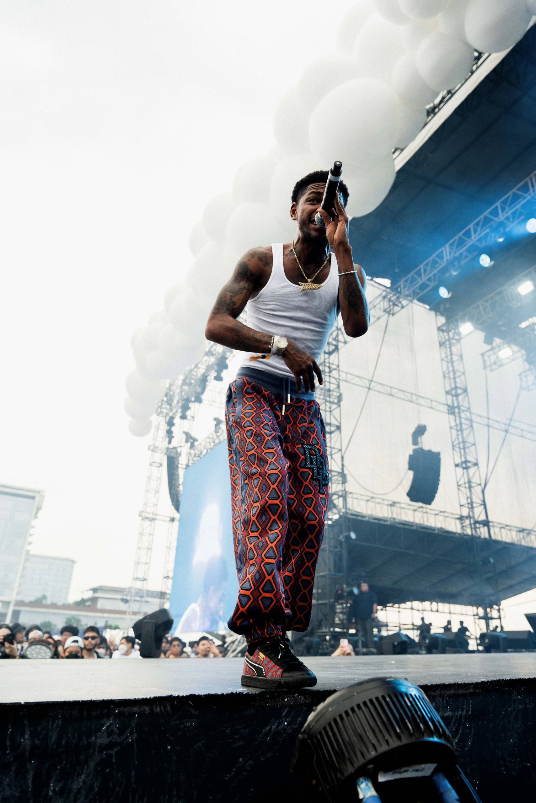 Rapper, musician Guap performs live on stage at a concert. He holds a mic in his hand as he wears a white tank top and red and orange-patterned pants.