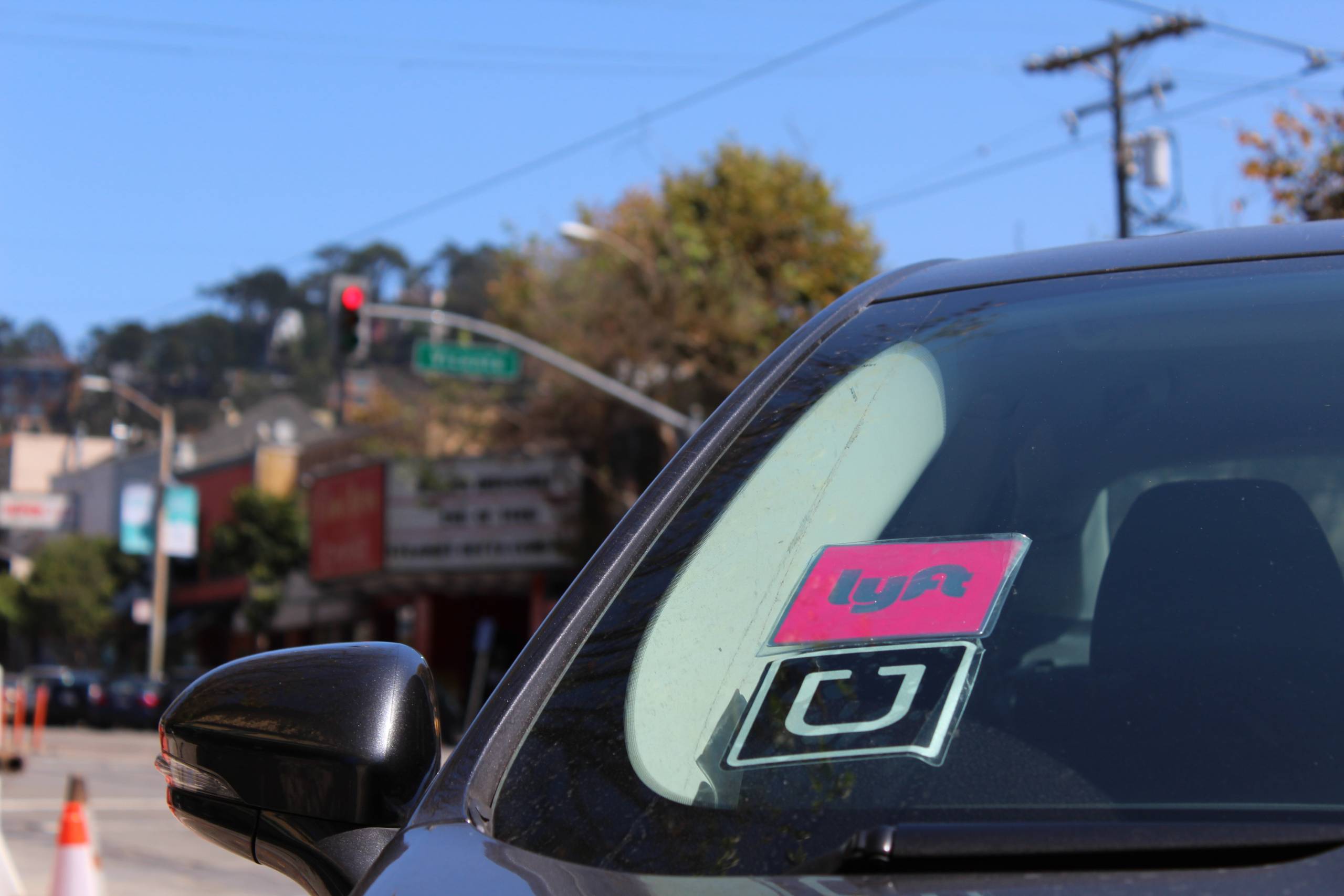 A closeup shot of a black vehicle with a pink Lyft sticker and a black and white Uber sticker on the left side of its windshield. The vehicle sits idle waiting to pick up a customer.