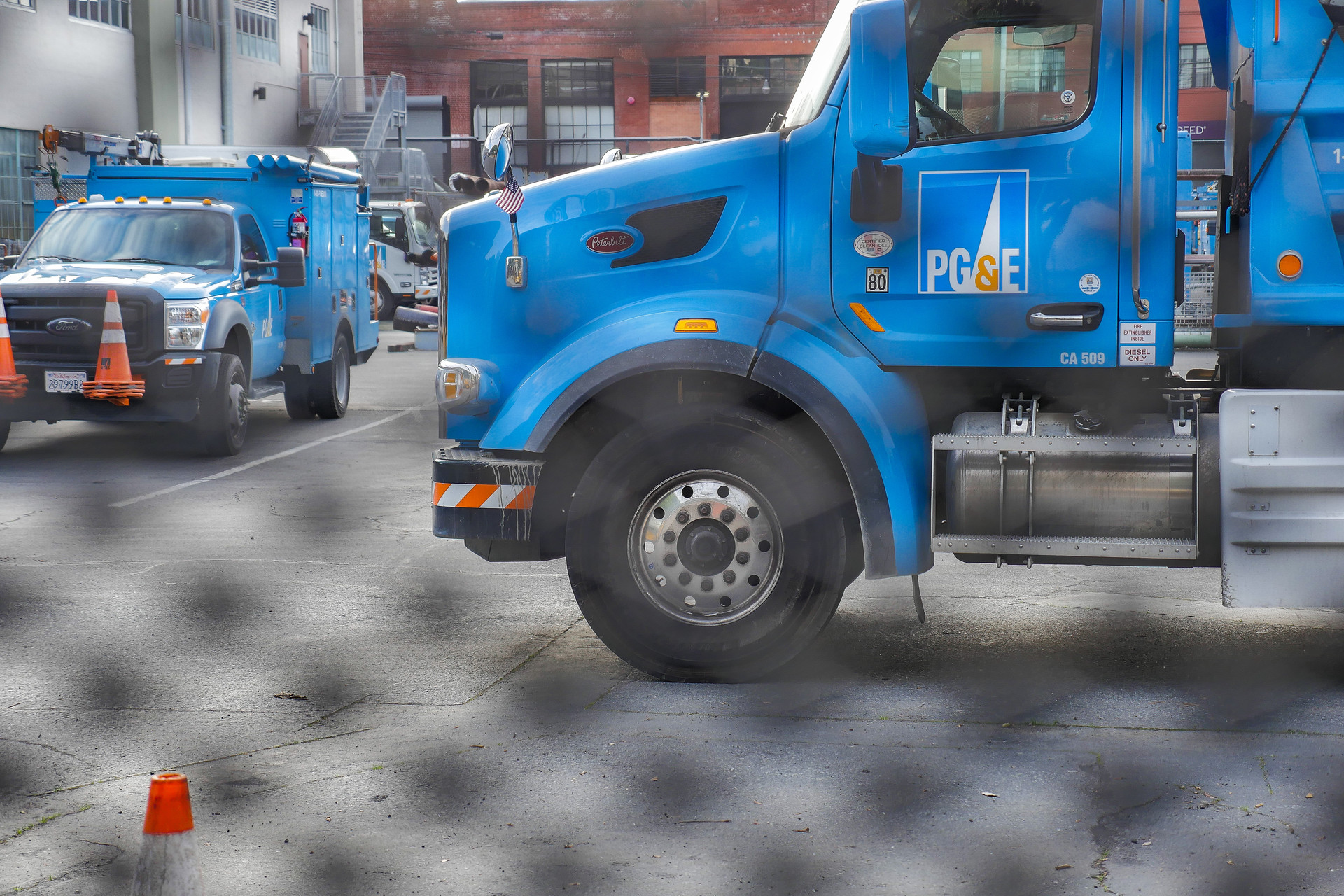 Blue trucks with 'pg and e' logo on them sit parked in a lot with the white and black blurry pattern of a fence in the foreground