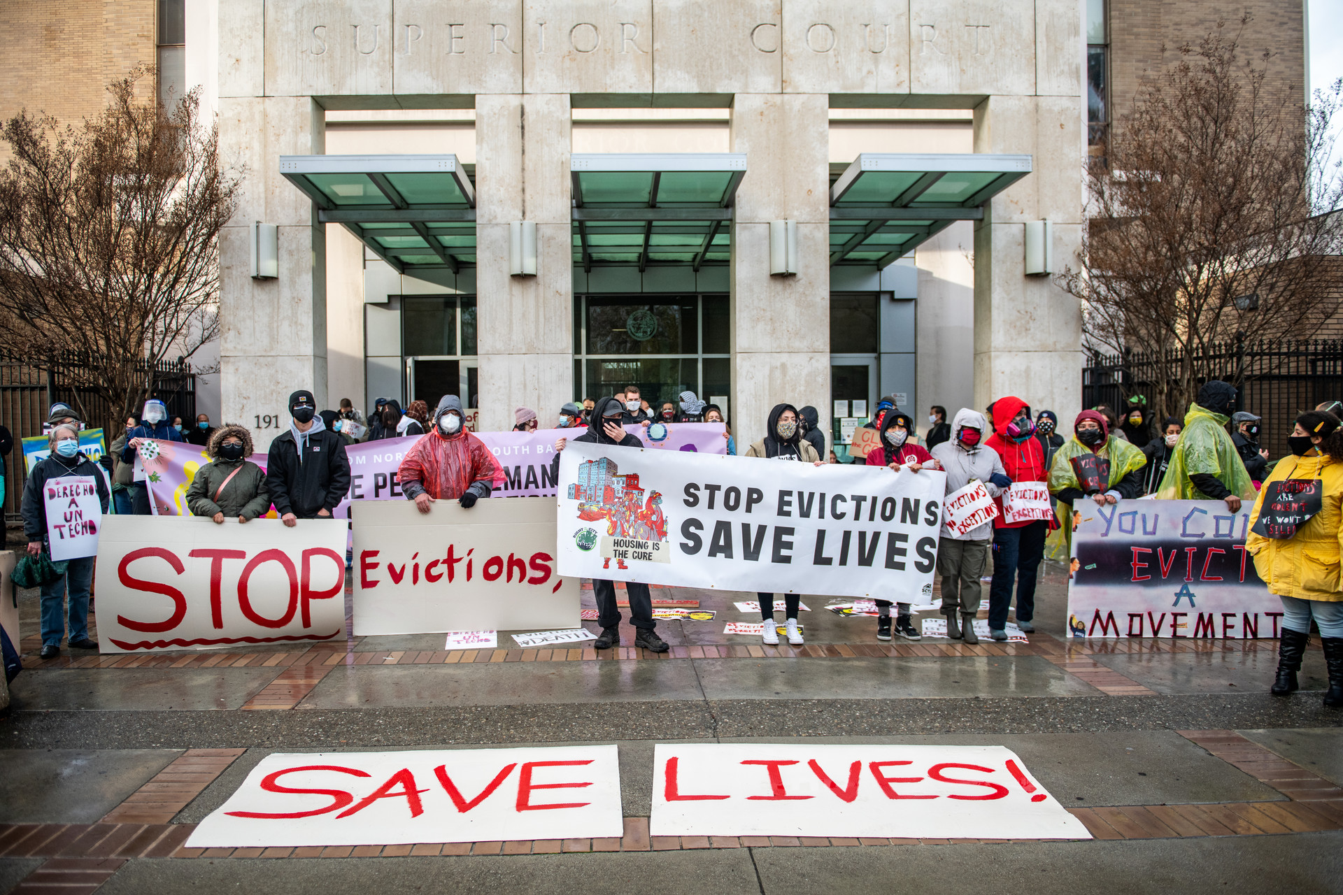 A wide shot of dozens of protestors in front of a superior court building. Large signs read "Stop Evictions, Save Lives."