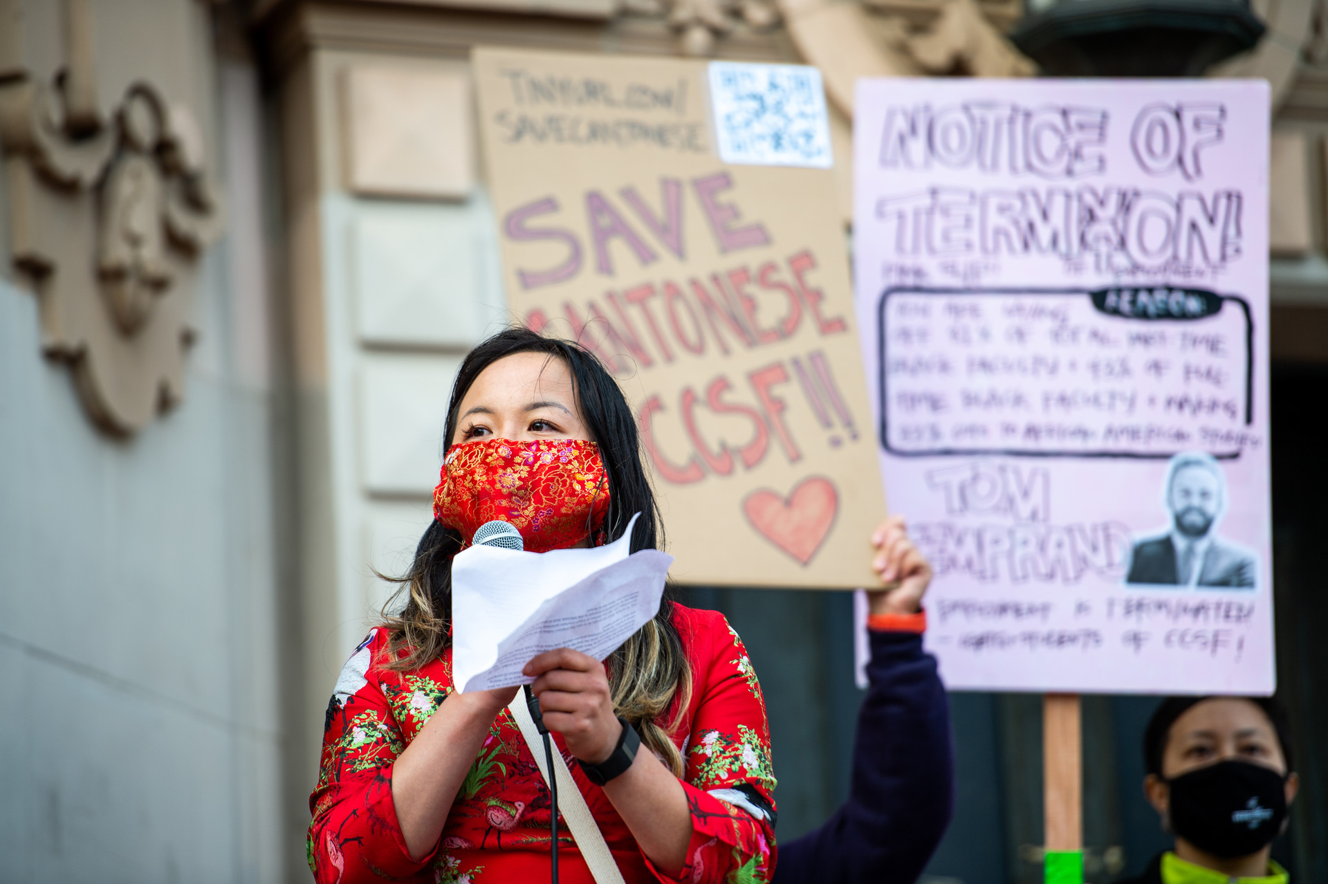 Asian woman wearing a colorful red shirt and a red-and-gold facemask holds a microphone with hand-made signs in the background reading Save Cantonese at CCSF