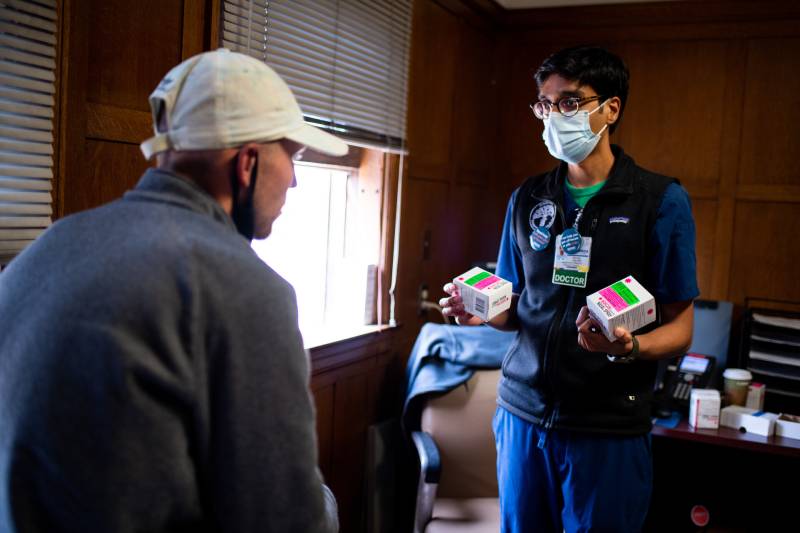 A medical professional in scrubs shows two boxes of the medicine Narcan to a patient wearing a white baseball cap.