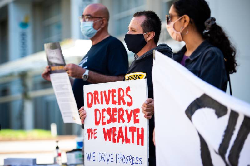 A man with dark hair holds a white sign with red lettering that reads, "Drivers Deserve the Wealth" and "We Drive Progress." Other black and white signs are seen in foreground.