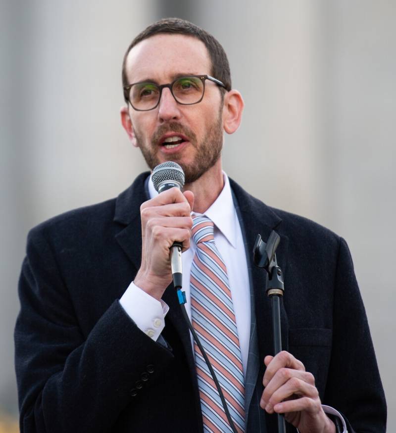 A white man in his 40s with glasses and a beard, wearing a black suit with a light colored diagonally striped tie talks into a microphone.