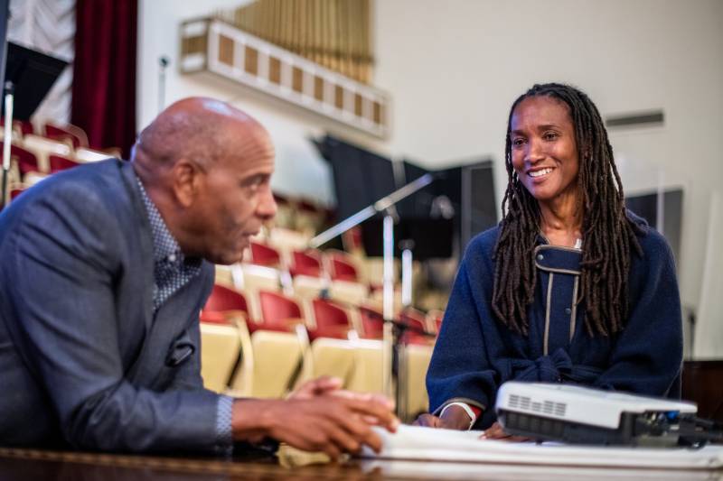 An African American woman smiles during a conversation with an African American man in a suit with empty seats in a conference hall in the background.