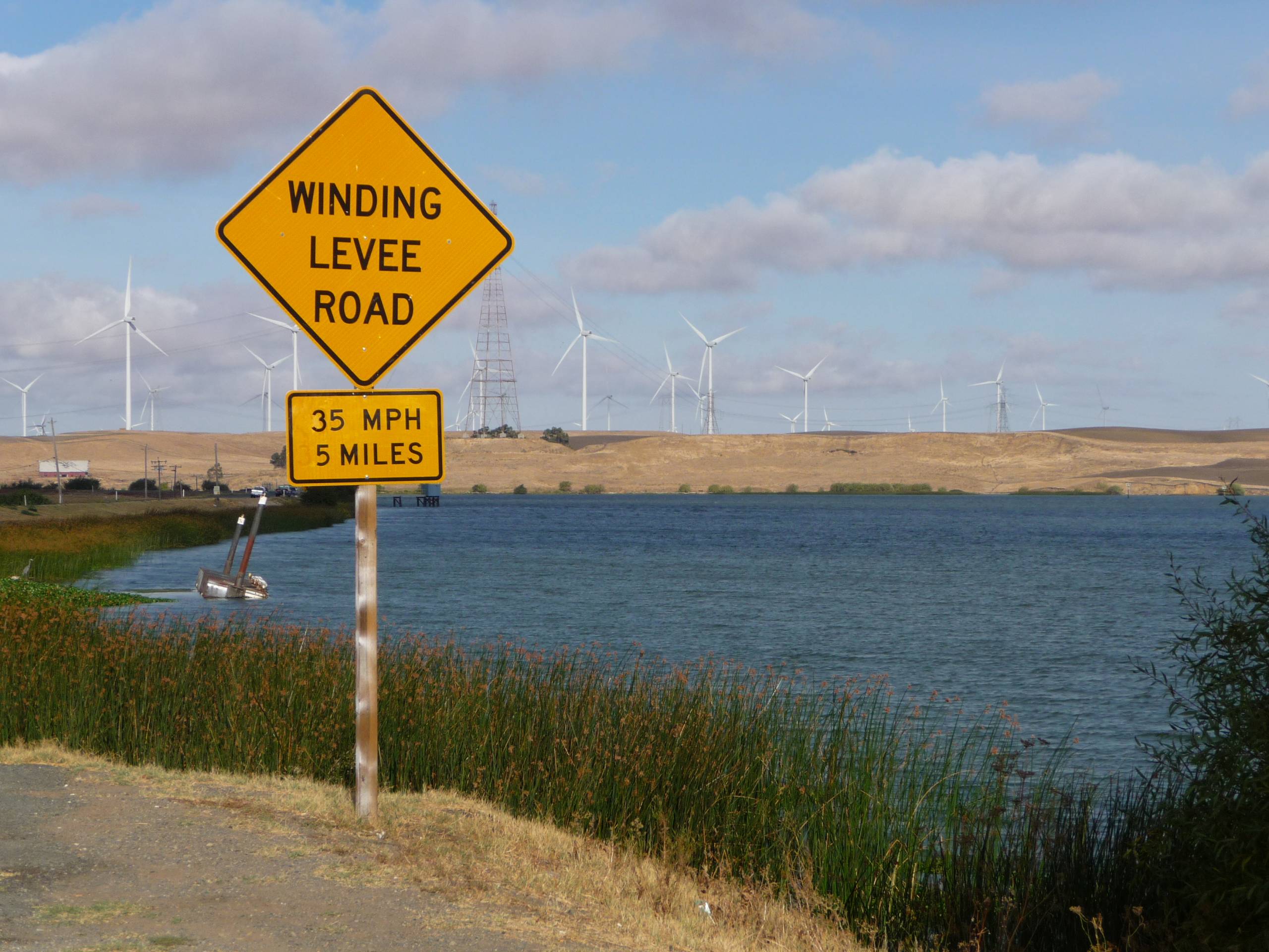 A diamond-shaped, yellow sign reads "Winding Levee Road." To the right of the sign, a body of water ripples as wind turbines spin in the background.