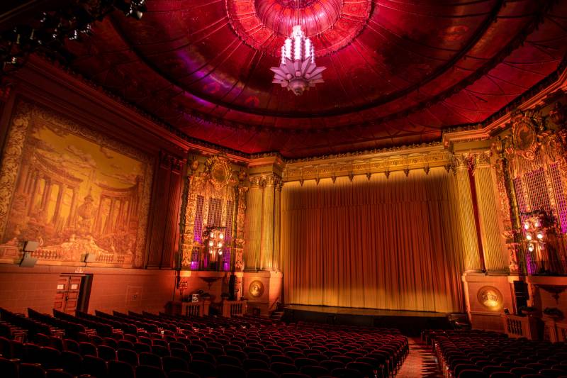 The inside of a grand movie theater with ornate decoration. An Art Deco chandelier hangs from a vaulted ceiling. Many rows of red velvet seats lead down to an elevated stage with a curtain closed over the film screen. The lighting is low, in reds and ambers.