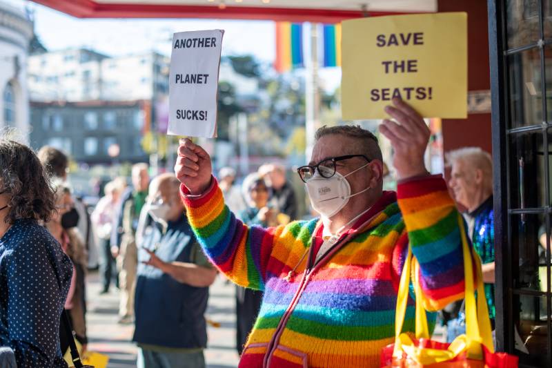 A person wearing a rainbow striped sweater hold up two signs. One says "Another Planet Sucks!" The other says "Save the seats."