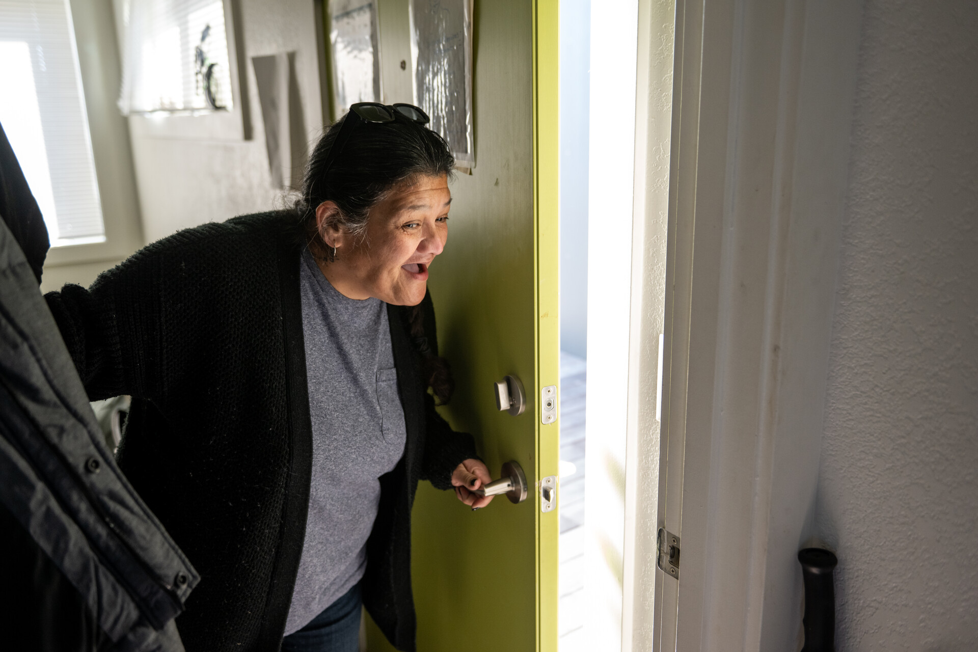 An older Latina woman, with short, graying black hair, wearing a gray T-shirt and a long-sleeved black cardigan, leans forward and smiles as she opens a bright yellow door.