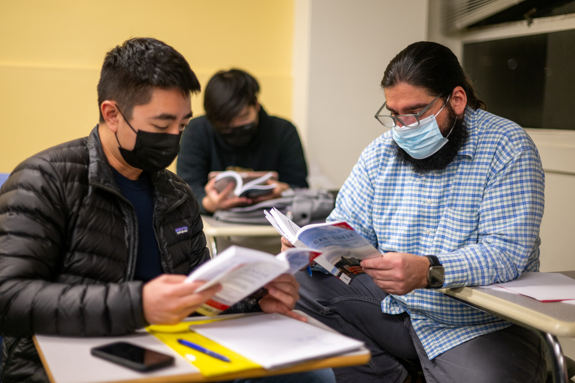 Two men wearing facemasks, one who appears to be Asian and one who appears to be white, face each other and hold school books while seated in a classroom setting