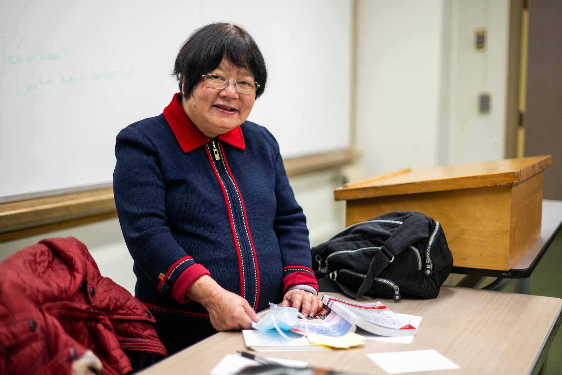 Middle-aged Asian woman wearing a blue and red zip-up top smiles as she stands behind a desk in a classroom with a white board behind her