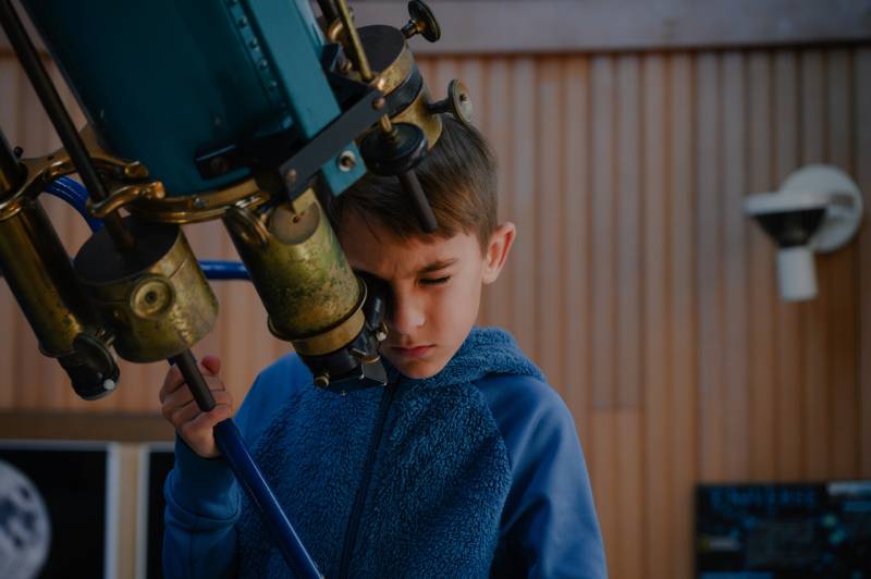 A young boy in a blue sweater closes one eye as he looks through a large telescope inside an observatory