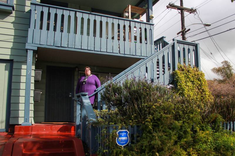 A woman wearing a purple shirt stands on stairs outside of a home.