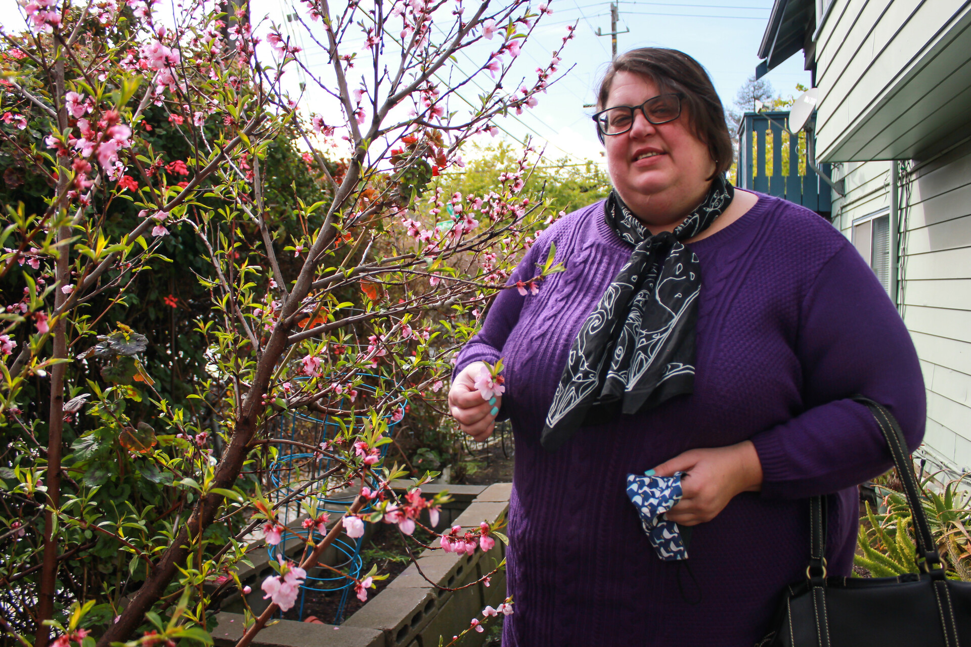 A large white woman with black-framed glasses and chin-length brown hair, wearing a purple sweater and black scarf knotted at the neck, with a black pocketbook crooked in her left elbow, stands smiling beside a pink, flowering plum branch on a wooden deck. She holds one of the pink blossoms pinched between finger and thumb in her right hand. 